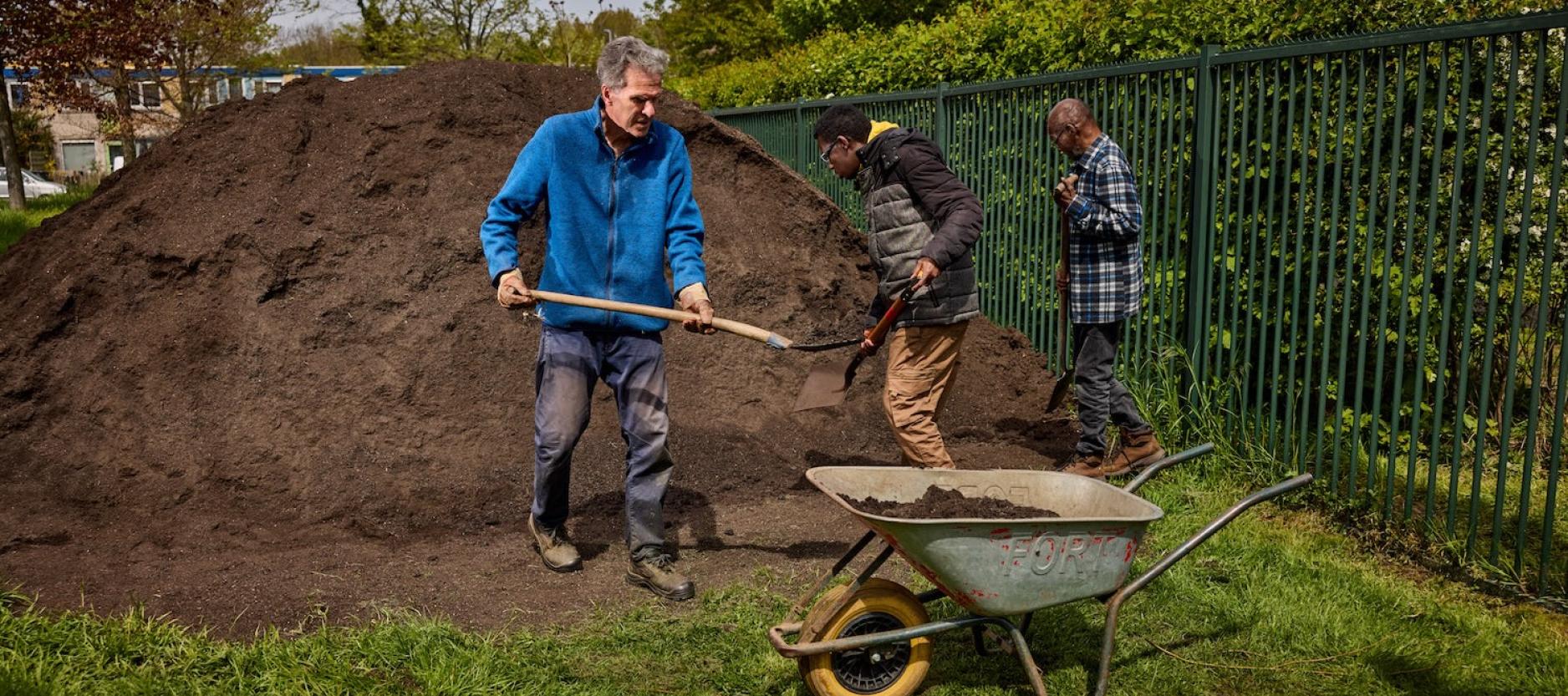 3 mannen met scheppen, bij een hoge aardeberg (1,5 meter) bezig met aarde in een kruiswagen aan het scheppen. 