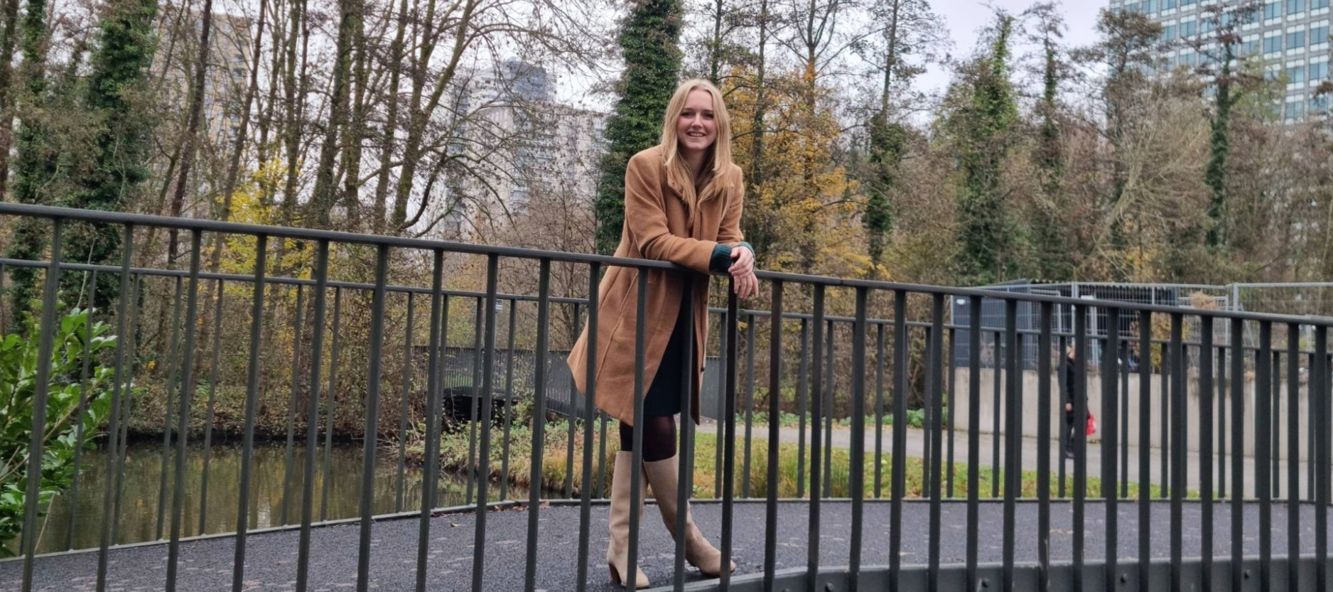 Een vrouw staat leunend op de leuning van een metalen voetgangersbrug in een parkachtige omgeving. Ze draagt een lange camel­kleurige jas en laarzen. Op de achtergrond zijn bomen met herfstkleuren en enkele hoge kantoorgebouwen te zien.